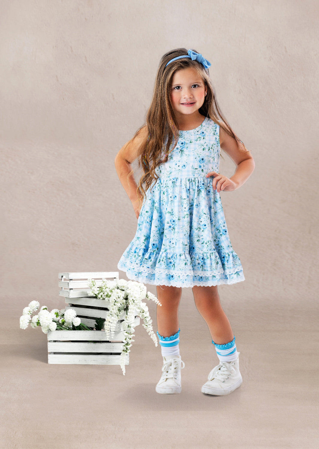 Young girl in a blue floral twirl dress standing next to wooden crates with flowers on a beige background