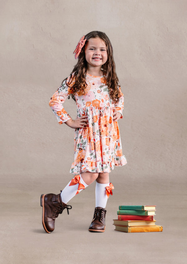 Toddler girl in a floral twirl dress standing next to stacked books on a beige background