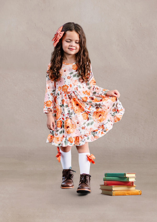 Young girl in a fall floral twirl dress standing next to stacked books against a plain background