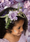Young girl wearing a floral headband with purple flowers and green leaves, surrounded by more flowers and balloons.