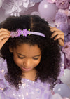Young girl wearing a purple headband with butterfly and floral decorations against a floral and balloon backdrop.