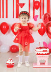 Child in a red dress celebrating a birthday with balloons and cake.