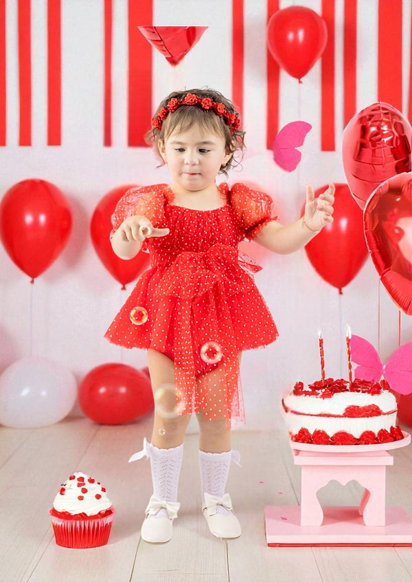 Child in a red dress celebrating a birthday with balloons and cake.