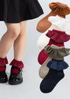 A child wearing black shoes with a burgundy ruffled cuff, shown beside a stack of children's socks in multiple colors with matching ruffled cuffs, on a light gray background.