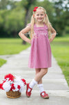 Little girl wearing a red and white gingham check dress with ruffle sleeves standing outdoors next to a basket of red and white pom-poms